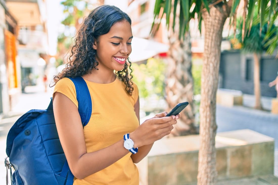 Jovem mulher sorrindo, em pé, olhando o celular